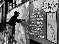 Sean works on his art piece next to a sign that reflects the usual topics in the Seattle area.